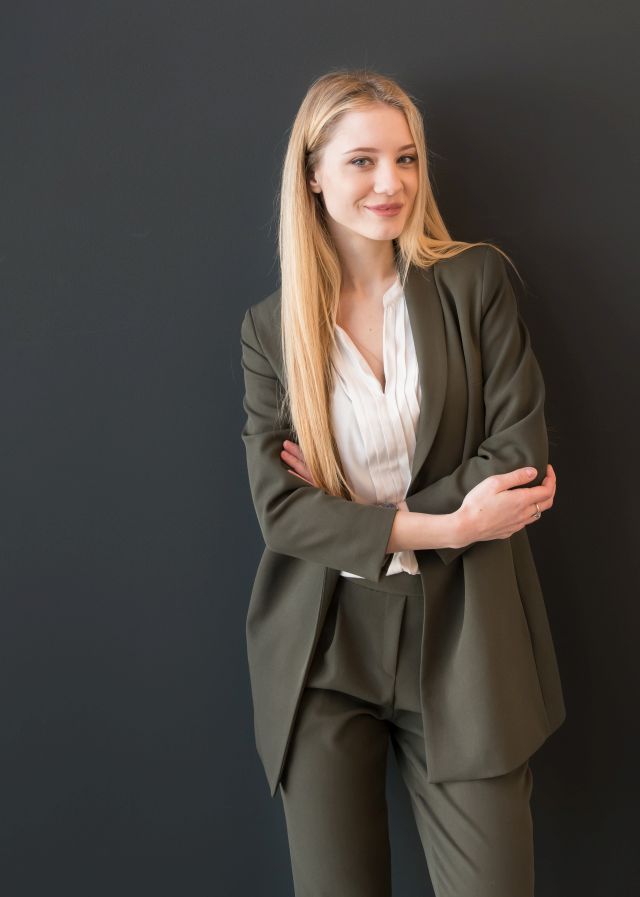 Beautiful blond young woman in dark elegant suit standing confidently with crossed arms and looking at camera smiling on black background